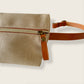 La riñonera Jud está realizada a mano en Barcelona con  tejido resistente al agua con una bonita cremallera dorada que le da un toque más chic. Disponible en 2 colores, gris oscuro y beige y puedes escoger la opción de la cinta en algodón o en piel.  Beige: disponible con cinta de algodón de espiga color natural con rayas blancas o cuero color camel.  Gris: Disponible con cinta algodón de espiga color beige oscuro o cuero color camel.  Puedes llevarla cruzada o como cinturón y puedes esconder el mosquetón e